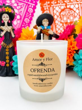 White glass Ofrenda soy candle with copal and sandalwood label before colorful Día de Muertos decorations.
