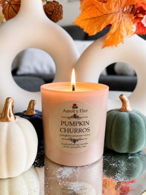 Lit pink glass Pumpkin Churros soy candle with pumpkin, cinnamon, and clove label among decorative pumpkins.