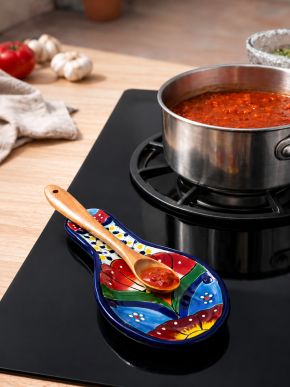 Painted ceramic spoon rest holding a salsa-covered wooden spoon beside a simmering saucepan.