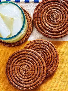 Overhead view of four handwoven pine needle coasters with a blue-rimmed drink on one.