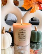 Lit pink glass Pumpkin Churros soy candle with pumpkin, cinnamon, and clove label among decorative pumpkins.